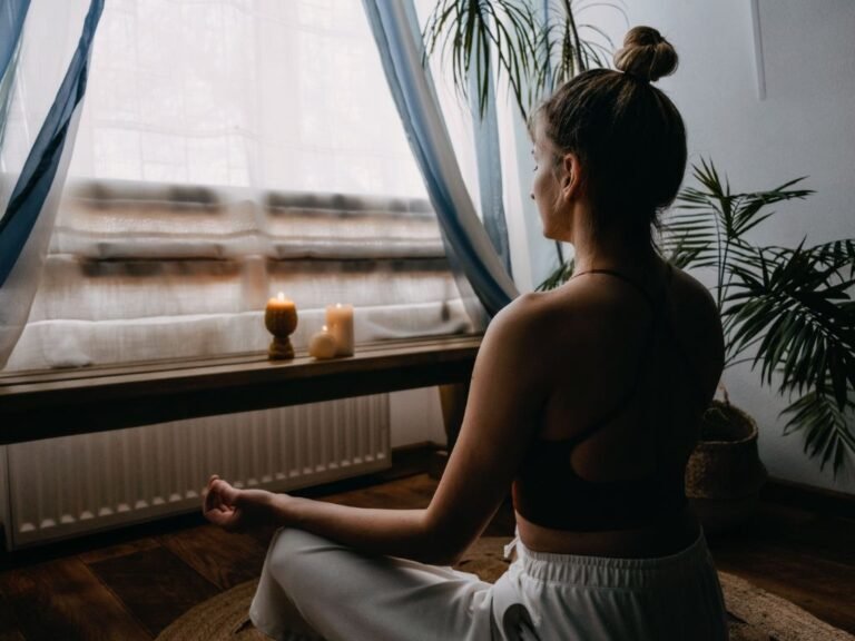 Woman meditating by a sunlit window with soft curtains.